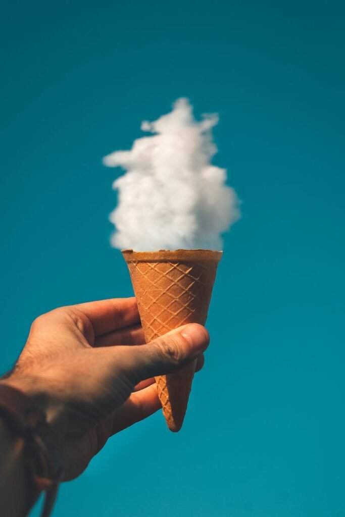 Hand holding an ice cream cone with a cloud effect against a clear sky background.