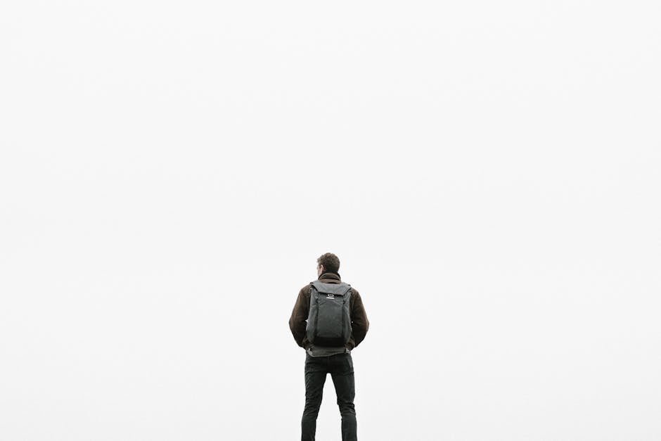 A man with a backpack stands alone facing a blank white background.